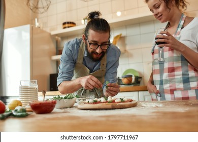 Best Cook. Young Couple Making Pizza Together At Home. Man In Apron, Professional Cook Adding Basil On The Dough While Woman Drinking Wine. Hobby, Lifestyle. Selective Focus. Horizontal Shot