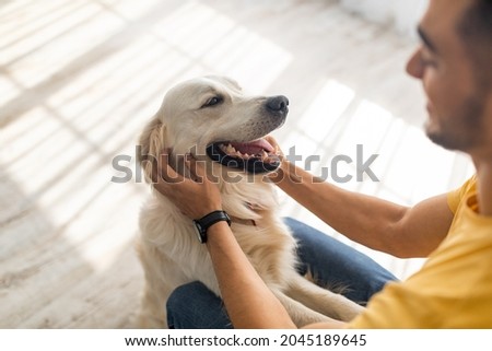 Best friend concept. Millennial Arab man scratching his dog on floor at home, selective focus. Young Eastern guy playing with his adorable pet, spending time with golden retriever indoors
