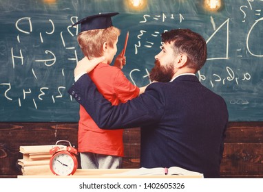 Best friends concept. Teacher with beard, father hugs little son in classroom while discussing, chalkboard on background. Child in graduate cap listening teacher, chalkboard on background, rear view.