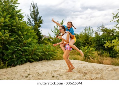 Best Friends Have Fun At The Beach In A Summer Park. Piggyback. Teen Girl Dressed In Shorts And A Shirt. On Summer Vacation. The Concept Of True Friendship.