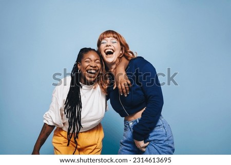 Best friends laughing and having a good time together in a studio. Happy young women enjoying themselves while standing against a blue background. Two vibrant female friends making memories.
