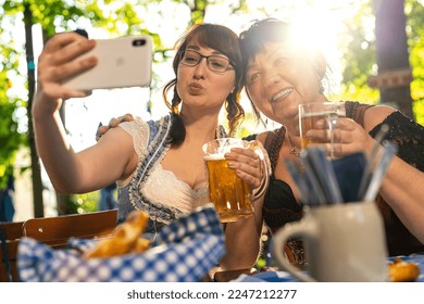 Best friends mother and daughter in Bavarian Tracht making a Selfie with the phone, with mugs of beer in Bavarian beer garden or oktoberfest - Powered by Shutterstock