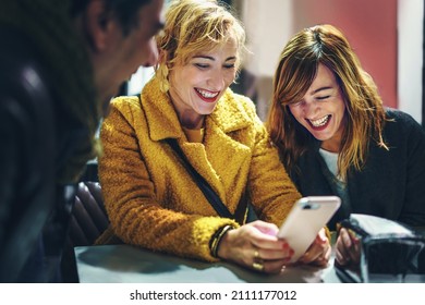 Best friends sitting at the coffee shop table watching funny meme or video streaming content on a digital mobile phone and laughing all together
