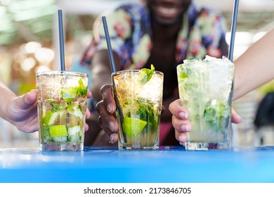 Best friends toasting cocktails. Multiethnic young people drinking long drinks at beach bar. Shallow depth of field with focus on hands holding iced mojitos. Close up shot, focus on the center glass. - Powered by Shutterstock