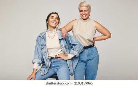 Best friends wearing denim jeans in a studio. Two happy women smiling at the camera cheerfully. Fashionable female friends posing together against a grey background.