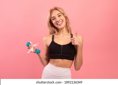 Best gift for active millennials. Fit blonde woman holding dumbbell with ribbon and showing thumb up gesture on pink studio background. Gym or sports club membership as present - Shutterstock ID 1892136349