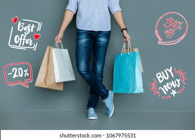Best Offer. Calm Young Man Standing Alone And Holding Bright Paper Bags After Buying Clothes At The Low Price