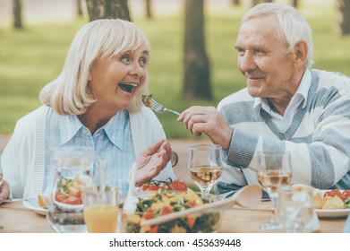 The Best For Our Nearest. Senior Man Feeding His Cheerful Wife With Fresh Salad While Both Sitting At The Dining Table Outdoors