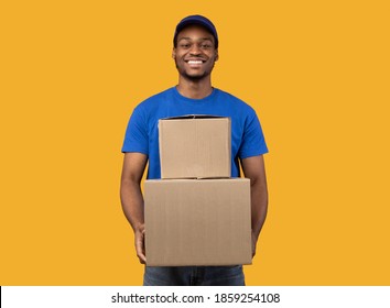 Best Service Concept. Smiling Black Delivery Man In Blue Cap And Unform Holding Pile Of Cardboard Boxes Standing Isolated Over Orange Studio Background. African American Courier Carrying Packages