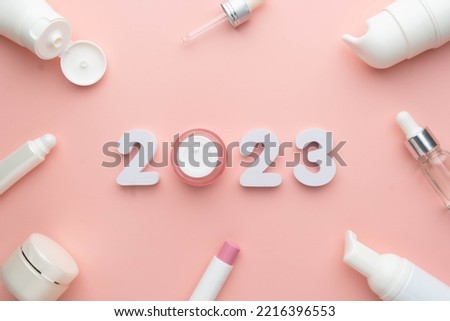 Best skincare products and cosmetic trends of 2023 concept. 2023 white number with lip balm, cream bottle, serum and lotion on pink background.