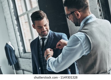 Best suit. Young fashionable designer helping his client to get dressed while standing in his workshop         - Powered by Shutterstock