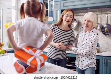 Best Surprise. Charming Young Woman And Her Senior Mother Trying To Guess What Kind Of Present Their Little Daughter Hiding Behind Her Back While Sitting On The Counter