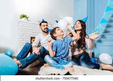 Best Time Together. Happy Young Family Sitting On The Floor On The White Rug And Throwing Balloons Into The Air While All Of Them Wearing Party Hats