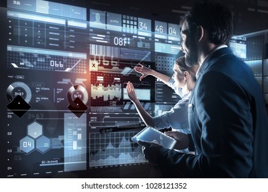 Best variant. Clever excited qualified programmers standing in front of a modern transparent screen and looking attentively at it while discussing their new amazing program - Powered by Shutterstock