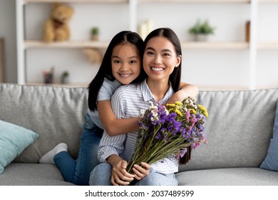 Best Wishes On Women Day. Happy Asian Girl Greeting Mom With Birthday Or Mother's Day, Giving Her Flowers And Embracing, Sitting Together On Sofa And Smiling To Camera