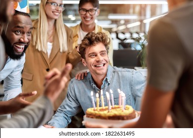 Best wishes. Young happy man blowing candles on cake while celebrating birthday among smiling mixed race business people. Office life. Corporate party - Powered by Shutterstock