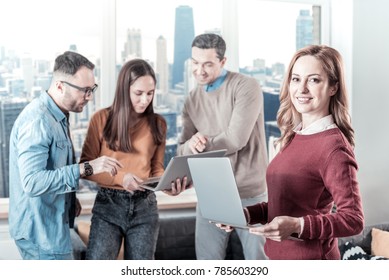 Best work. Confident pretty beautiful woman standing near her colleagues holding the laptop and looking straight.