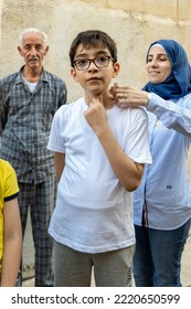 Bethlehem, West Bank, Palestine - 22 July 2022: Young Arab Boy With White Shirt And Black Glasses Thoughtfully Stares At The Lense With Old Man And Woman With Blue Hijab In The Background Grabbing Him