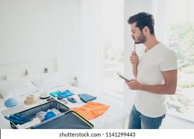 It is better to write everything! Young bearded man standing in a bright bedroom in front of clothes thinks what else needs to be folded into a suitcase before traveling - Powered by Shutterstock