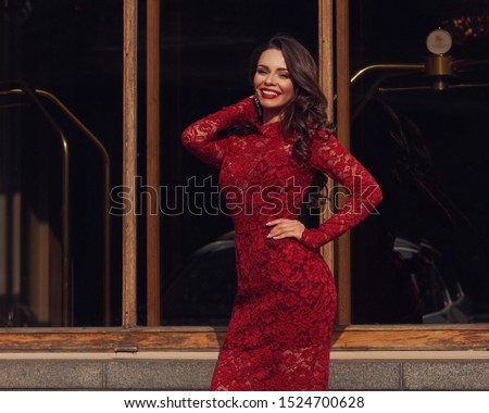Beuatiful elegant woman wearing red lace dress. Lady with makep and wvy brunette hair standing and posing against wall with window on a sunny day