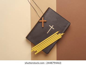 Bible book, candles and wooden Christian cross on a string, beige background. Religion, prayer, going to church concept. Top view. Flat l;ay