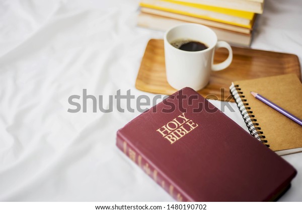 ้Holy bible with small note book,\
 pencil and a cup of black coffee over blurred book stack on bed,\
Christian background morning devotional concept with copy\
space