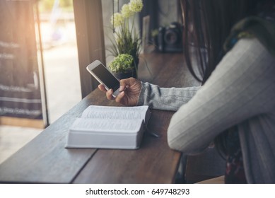 Bible Study Concept. Sad  Christian Woman Read Bible At Coffee Shop, Send Message By Mobile Phone. Person Side View, Hold Smartphone. Asian Student Reading Book And Study For Exam In Library.