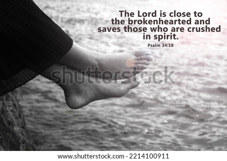 Bible verse quote - The Lord is close to the brokenhearted and saves those who are crushed in spirit. Psalm 34:38 with young woman sitting alone with bare feet on the water in black and white.