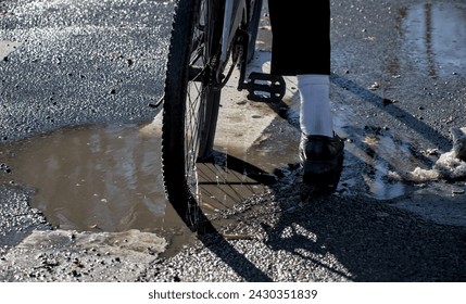 bicycle rear wheel in deep pothole filled with dirty water during winter (road construction, bike stuck inside hole) street streets crosswalk (shoe sock pants) detail close up bad conditions - Powered by Shutterstock