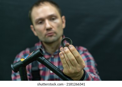 Bicycle Repair. The Bike Mechanic Changes The Cups And Bearings In The Headset Of The Frame. Bicycle Parts In The Workshop On A Black Background. The Face Of A Young Guy With A Tool In His Hands.