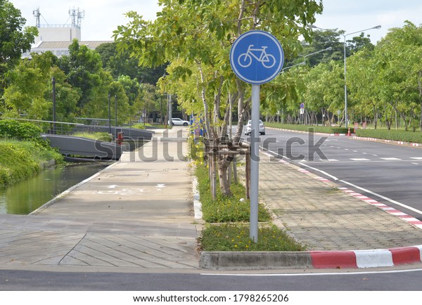 Bicycle riding sign in front of the bike\
path divided  from footpath with line of trees leading to the\
building with communication poles of the roof\
top.