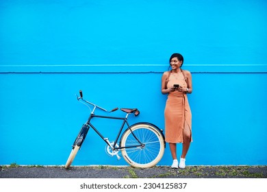 Bicicleta, joven mujer con smartphone y feliz afuera por la pared azul. Ciclismo en el área urbana, salud y bienestar y una persona femenina en el teléfono celular al aire libre parado en las calles con bicicleta o bicicleta en la carretera Foto de stock