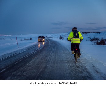A bicyclist in a reflective vest rides his bike on a icy country road during polar night twilight in January, in Norway, 500 km far to the North from Polar circle. Real life situation.
