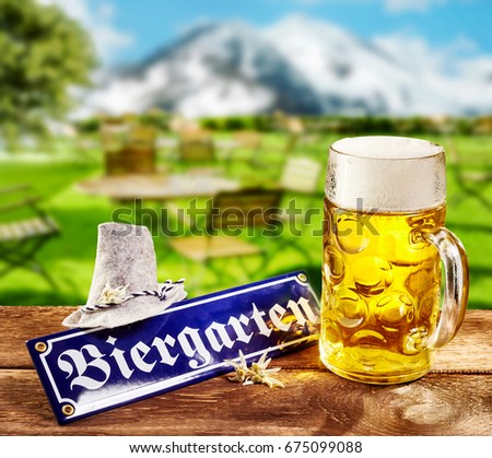 Biergarten or Beer Garden sign for German called Oktoberfest lying on a rustic outdoors wooden table with a full frothy glass mug of beer and hat with a background view of the Bavarian Alps