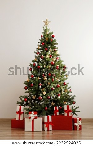 Big beautiful christmas tree decorated with shiny baubles and many different presents on wooden floor. White wall background with a lot of copy space for text. Close up.