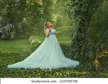 A Big, Beautiful, Young Woman In A Luxurious Blue Dress With A Long Train Is Walking In A Blooming Garden. The Model Size Is Plus. The Hair Is Large With Curls. Artistic Photo