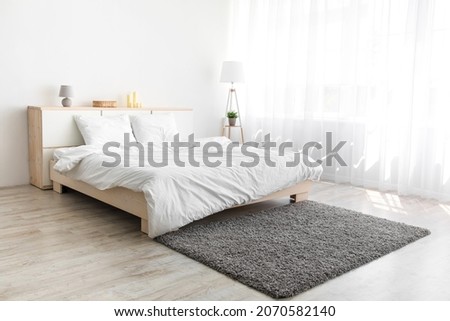 Big bed with comfortable bedding, soft gray carpet, lamp on floor, furniture with accessories, window with curtains, white wall. Scandinavian bedroom interior, mortgage, design blog and furniture ad