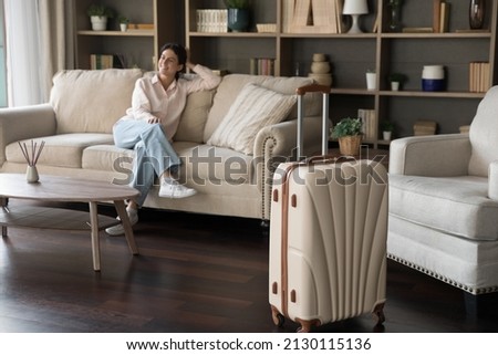 Big beige suitcase standing on wooden floor in modern hotel room with relaxed millennial Hispanic woman sitting on comfortable couch. Joyful lady feeling excited returning home after vacation travel.