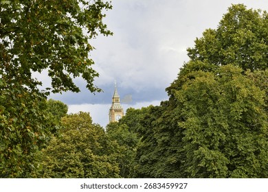 Big Ben clock tower peeks through dense, green foliage under a cloudy, overcast sky  in London, UK.