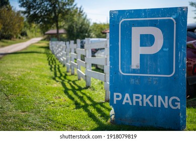 Big blue parking sign, leaning against old wooden white cattle fence, on a countryside road. A farm house in background.