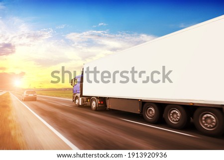 Big blue truck driving fast with a white trailer with blank space for text on a countryside road with other cars against a blue sky with a sunset