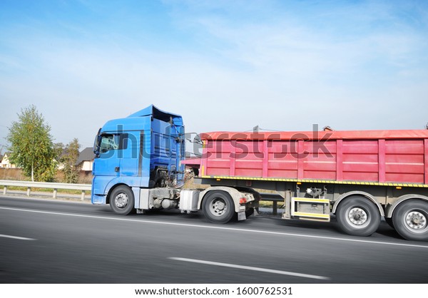 Big blue truck with\
red trailer in motion on the countryside road with houses against\
blue sky with clouds