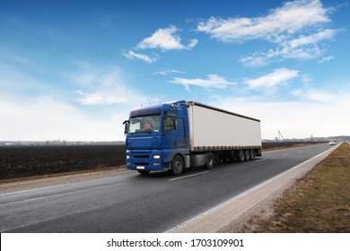 A big blue truck and a white trailer with space for text on the countryside road against a blue sky with clouds