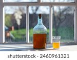 Big bottle with a drink made from fermented birch sap on the windowsill on a warm spring day, close up. Traditional Ukrainian cold barley drink kvass in a glass jar and glass on table near yard
