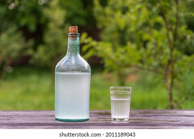 Big Bottle And Glass Of Moonshine Or Vodka On Wooden Table In Sunny Summer Day In Garden, Close Up. Traditional Ukrainian Alcoholic Drink