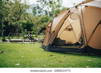 A big brown dome camping tent in a green grass with trees and mountains. Taken in an afternoon with a clear blue sky with white clouds. There're chairs, table and camping equipments nearby. - Powered by Shutterstock