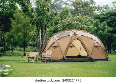 A big brown dome camping tent in a green grass with trees and mountains. Taken in an afternoon with a clear blue sky with white clouds. There're chairs, table and camping equipments nearby. - Powered by Shutterstock