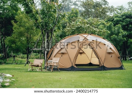 A big brown dome camping tent in a green grass with trees and mountains. Taken in an afternoon with a clear blue sky with white clouds. There're chairs, table and camping equipments nearby.