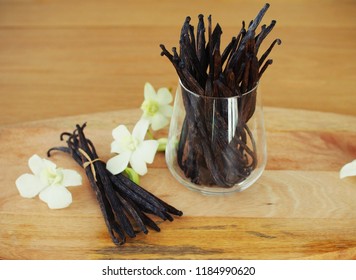 A big bunch of vanilla beans in the glass and a smaller on on the wooden board with white orchids