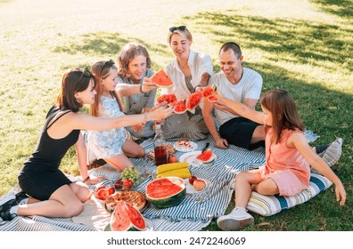 Big caucasian family on picnic blanket on the in city park green grass. They rose-up red juicy watermelon pieces and clinking them like glasses. Family values and outdoor activities concept. - Powered by Shutterstock